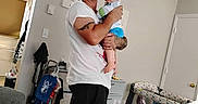 James is registered to the contest to win money with this photo: adult, baby, child, father, holding, indoors, living_room, nursery, parenting, portrait, standing, smiling, cap, tattoo, white_shirt, playpen, crib, stuffed_animal, furniture, family