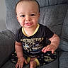 James is registered to the contest to win money with this photo: baby, child, infant, sitting, armchair, portrait, smiling, face, eyes, teeth, short_hair, black_shirt, patterned_clothing, construction_theme, toy_vehicles, indoor, furniture, gray_background, photography, happy