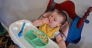 James is registered to the contest to win money with this photo: baby, bib, chair, child, eating, face, feeding, food, hands, high_chair, home, indoor, kitchen, plate, portrait, sitting, snack, spoon, wooden_furniture, yellow_clothing