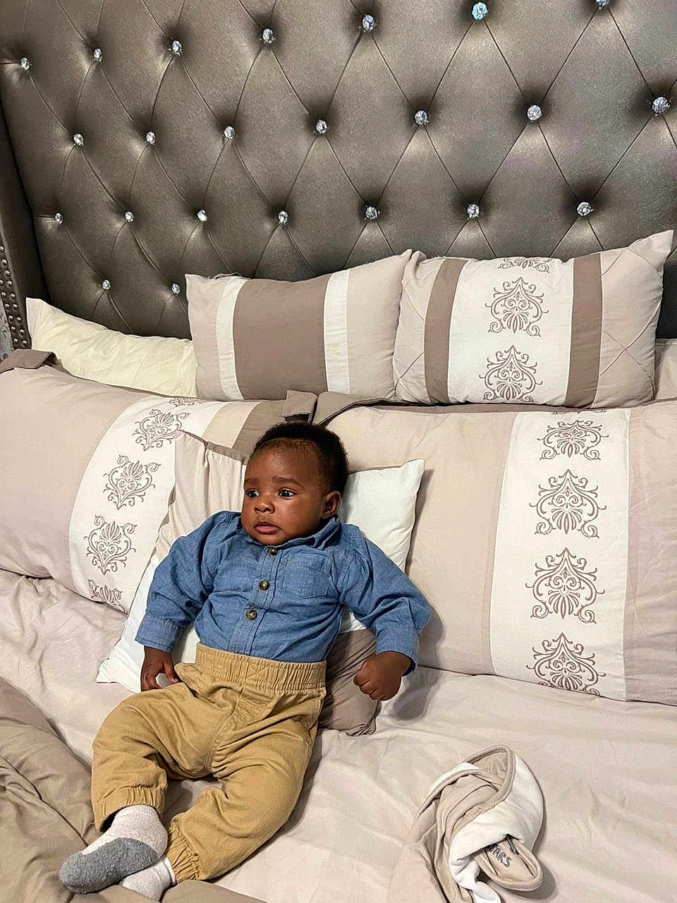 Liam is registered to the contest to win money with this photo: baby, child, bed, pillow, headboard, blue_shirt, beige_pants, socks, curious, indoor, furniture, decorative_pillows, tufted_headboard, soft_lighting, relaxed, infant, casual_clothing, cozy, home, portrait