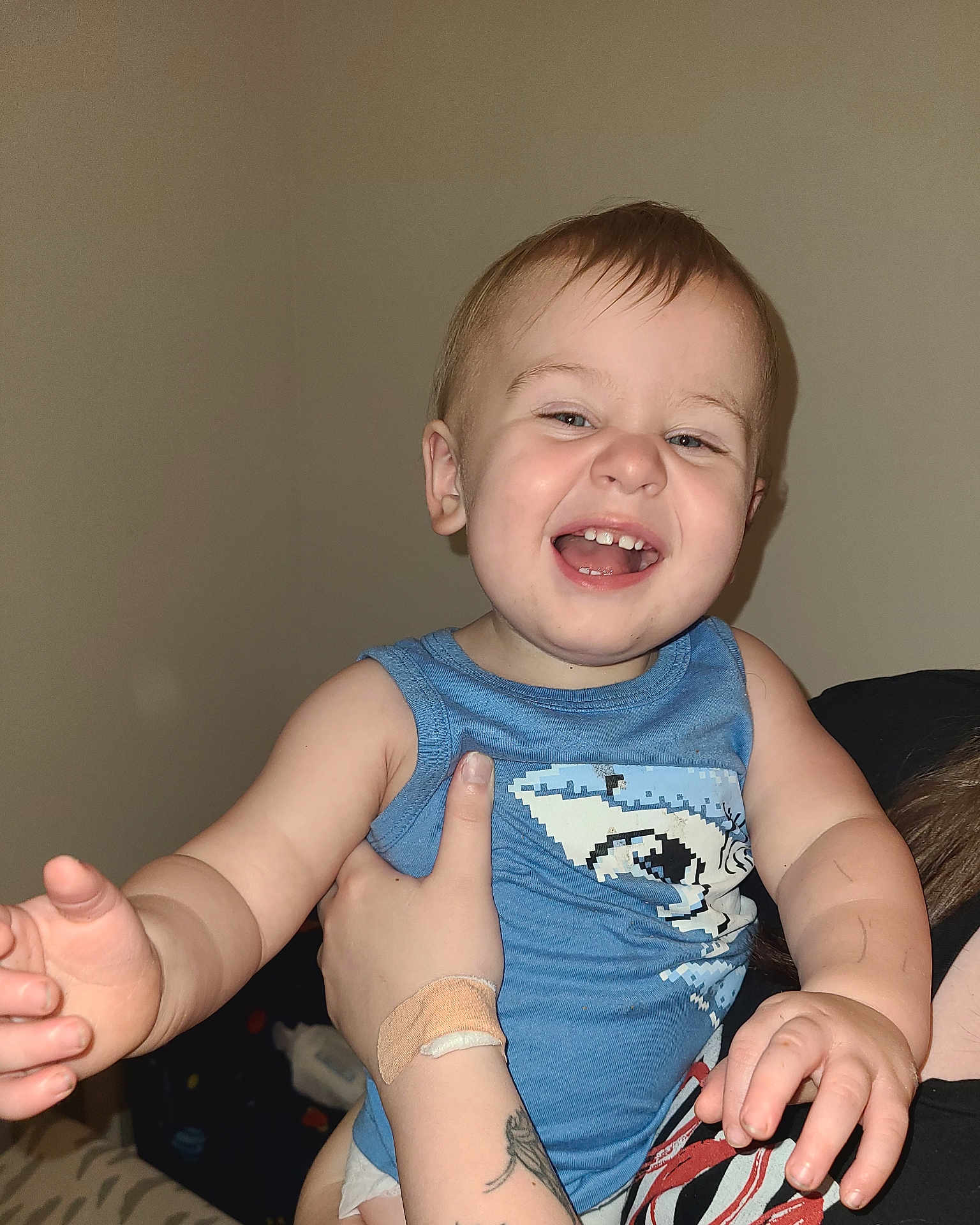 Jacob is registered to the contest to win money with this photo: toddler, child, smiling, happy, blue_shirt, pixelated_shark, indoor, person, hand, arm, skin, face, hair, diaper, tattoo, bandage, adult_hand, holding, baby, cute