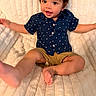 Benjamin is registered to the contest to win money with this photo: baby, child, infant, curly_hair, smiling, sitting, portrait, indoor, blanket, quilt, bed, navy_shirt, star_pattern, shorts, barefoot, arms_outstretched, happy, cozy, close_up, photography