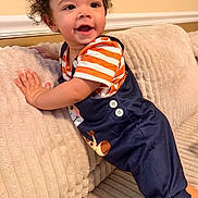 Benjamin is registered to the contest to win money with this photo: toddler, child, baby, curly_hair, smiling, happy, sitting, couch, sofa, pillow, blanket, portrait, indoor, home, orange_shirt, striped_shirt, navy_overalls, cozy, cute, face