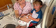 Jolene joined the competition — help win amazing prizes! woman, child, baby, hospital_bed, pajamas, blanket, pillow, hospital_room, medical_equipment, remote_control, cup, table, wooden_cabinet, curtains, smiling, holding, sitting, person, newborn, family