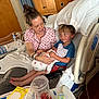 woman, child, baby, hospital_bed, pajamas, blanket, pillow, hospital_room, medical_equipment, remote_control, cup, table, wooden_cabinet, curtains, smiling, holding, sitting, person, newborn, family