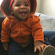 Malik is registered to the contest to win money with this photo: baby, child, infant, smiling, happy, portrait, orange_jacket, sun_hat, winter_clothing, shoe, sitting, couch, indoors, face, person, outdoor_style, cozy, fashion, playful, closeup