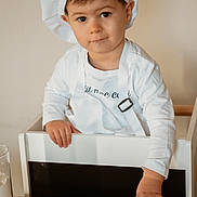 Noah participe au concours pour gagner de l'argent avec cette photo : child, toddler, chef_hat, apron, rolling_pin, dough, flour, kitchen, table, high_chair, baking, cooking, wooden, person, cute, indoor, young_child, food_preparation, white_clothing, playful