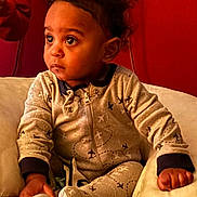 Kyrie is registered to the contest to win money with this photo: toddler, child, baby, onesie, airplane_pattern, curly_hair, indoor, cushion, toy, seated, expression, face, hand, cozy, red_background, comfort, clothing, portrait, young_child, curiosity