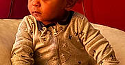 Kyrie is registered to the contest to win money with this photo: airplane_pattern, baby, child, clothing, comfort, cozy, curiosity, curly_hair, cushion, expression, face, hand, indoor, onesie, portrait, red_background, seated, toddler, toy, young_child