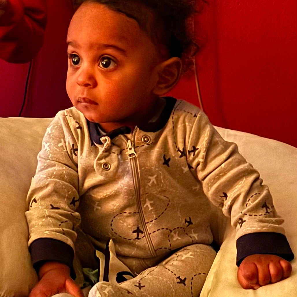 Kyrie is registered to the contest to win money with this photo: toddler, child, baby, onesie, airplane_pattern, curly_hair, indoor, cushion, toy, seated, expression, face, hand, cozy, red_background, comfort, clothing, portrait, young_child, curiosity