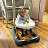 Kyrie is registered to the contest to win money with this photo: toddler, baby_walker, child, indoor, wooden_floor, table, chair, snack, plate, sweater, black_socks, broom, dustpan, kitchen, wall, flooring, furniture, curious, seated, looking_up