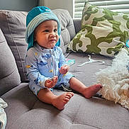Jayceon is registered to the contest to win money with this photo: baby, child, blue_hat, onesie, sitting, couch, gray_couch, pillow, green_pillow, indoor, window_blinds, curious_expression, barefoot, small_object, cozy, home, furniture, infant, cute, casual