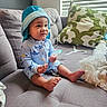 baby, child, blue_hat, onesie, sitting, couch, gray_couch, pillow, green_pillow, indoor, window_blinds, curious_expression, barefoot, small_object, cozy, home, furniture, infant, cute, casual
