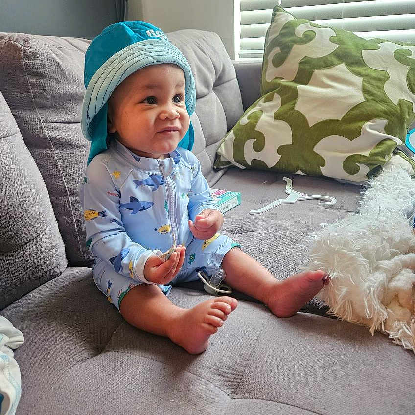 Jayceon is registered to the contest to win money with this photo: baby, child, blue_hat, onesie, sitting, couch, gray_couch, pillow, green_pillow, indoor, window_blinds, curious_expression, barefoot, small_object, cozy, home, furniture, infant, cute, casual