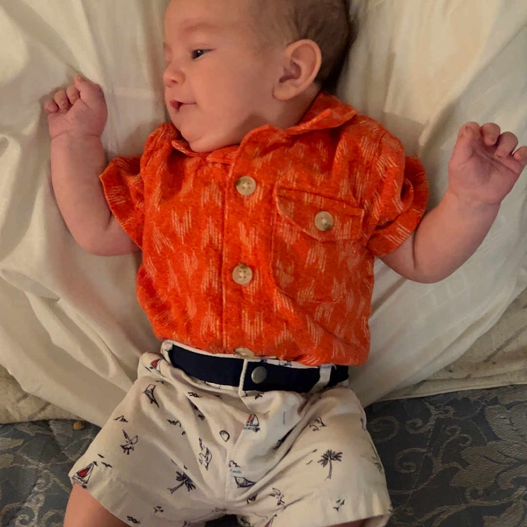 Johnathon Reinhardt is registered to the contest to win money with this photo: baby, bed, child, clothing, cute, face, feet, hands, indoor, infant, lying_down, mattress, orange_shirt, patterned, pillow, portrait, relaxed, shorts, skin, small