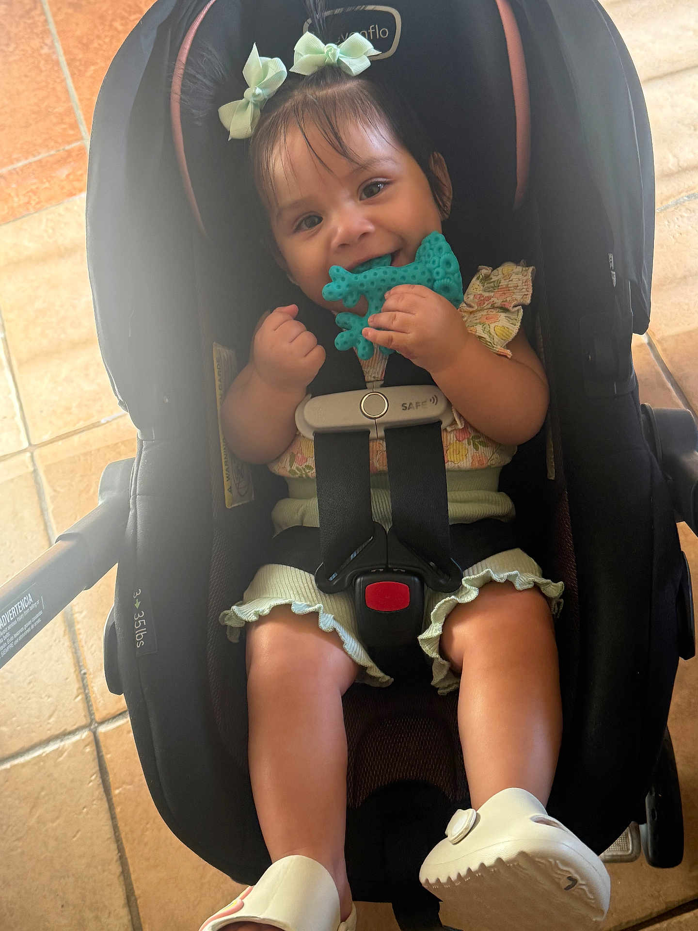 Maria joined the competition — help win amazing prizes! baby, child, car_seat, teething_toy, smiling, shoes, bows, clothing, sitting, indoor, floor, tile, straps, happy, person, cute, infant, chewing, play, secure