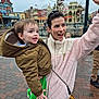 buildings, child, daytime, green_clothing, hand_raised, happy, jacket, mouse_ears, outdoor, pavement, people, polka_dots, portrait, red_skirt, smiling, theme_park, toddler, tree, winter_clothing, woman