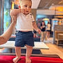 Monté a rejoint le concours — aidez-le/la à gagner de superbes lots ! baby, child, indoor, cafe, polo_shirt, shorts, barefoot, red_couch, hand, person, furniture, table, blurred_background, lighting, curious, standing, casual_clothing, young_child, holding, portrait