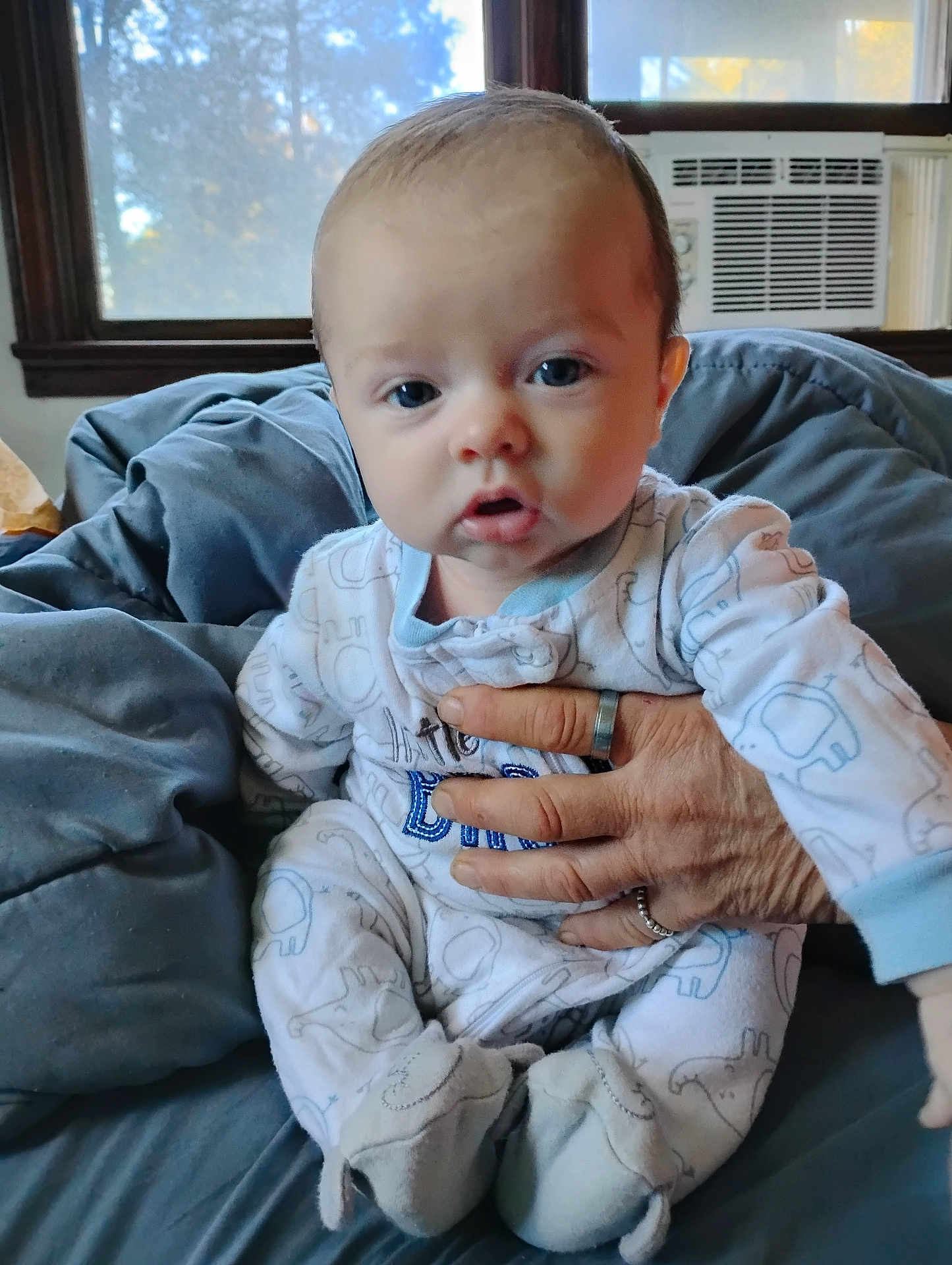Kiah joined the competition — help win amazing prizes! baby, infant, pajamas, elephant_pattern, hand, adult_hand, blanket, window, air_conditioner, indoor, portrait, cute, child, soft_toys, footed_pajamas, blue, white, face, expression, cozy