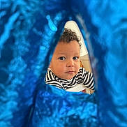 Anès a rejoint le concours — aidez-le/la à gagner de superbes lots ! toddler, child, face, curly_hair, striped_shirt, balloon, blue, foil, peek, curious, indoor, portrait, expression, young_child, closeup, person, cute, playful, happy, background_blur
