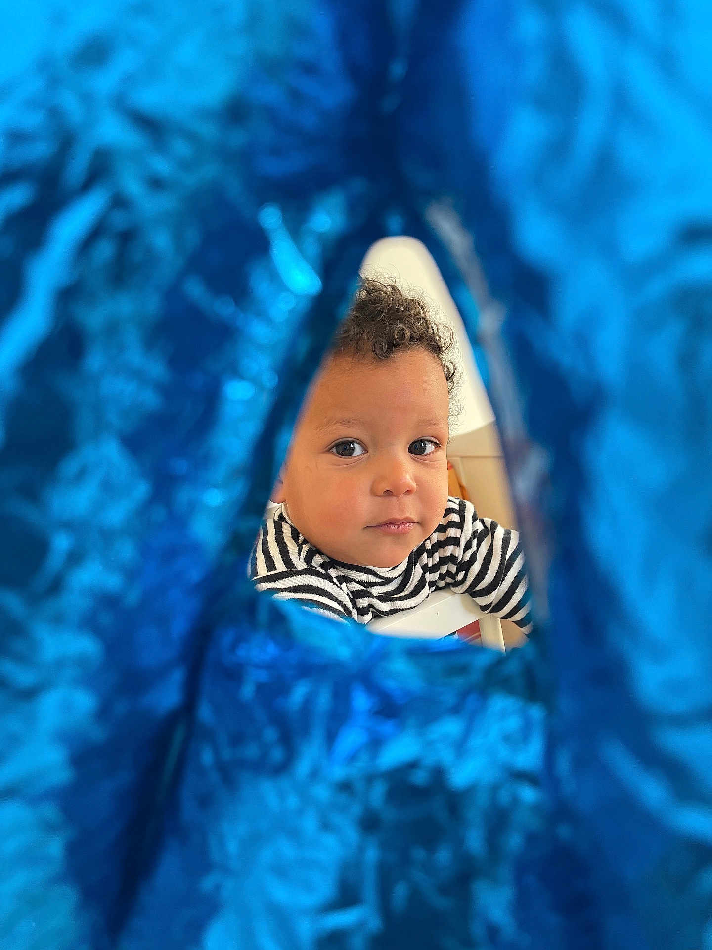 Anès a rejoint le concours — aidez-le/la à gagner de superbes lots ! toddler, child, face, curly_hair, striped_shirt, balloon, blue, foil, peek, curious, indoor, portrait, expression, young_child, closeup, person, cute, playful, happy, background_blur