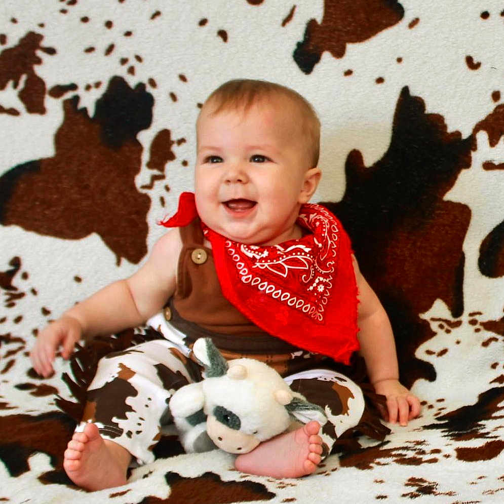 William is registered to the contest to win money with this photo: baby, blanket, brown, child, clothing, cow_pattern, cute, face, foot, happy, indoor, infant, person, plush_toy, portrait, red_bandana, sitting, smiling, toy, white