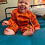 baby, bed, blue_bedspread, child, cute, foot, framed_picture, hand, head, indoor, infant, orange_clothing, person, playful, portrait, pumpkin_pattern, sitting, smiling, tongue_out, wall_art