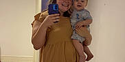 William is registered to the contest to win money with this photo: baby, ceiling, chandelier, changing_table, dinosaur_print, doorway, dress, floor_tiles, handheld_phone, happy, holding_baby, indoor, mirror_selfie, pajamas, person, reflection, room, smile, wall, woman