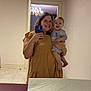 baby, ceiling, chandelier, changing_table, dinosaur_print, doorway, dress, floor_tiles, handheld_phone, happy, holding_baby, indoor, mirror_selfie, pajamas, person, reflection, room, smile, wall, woman