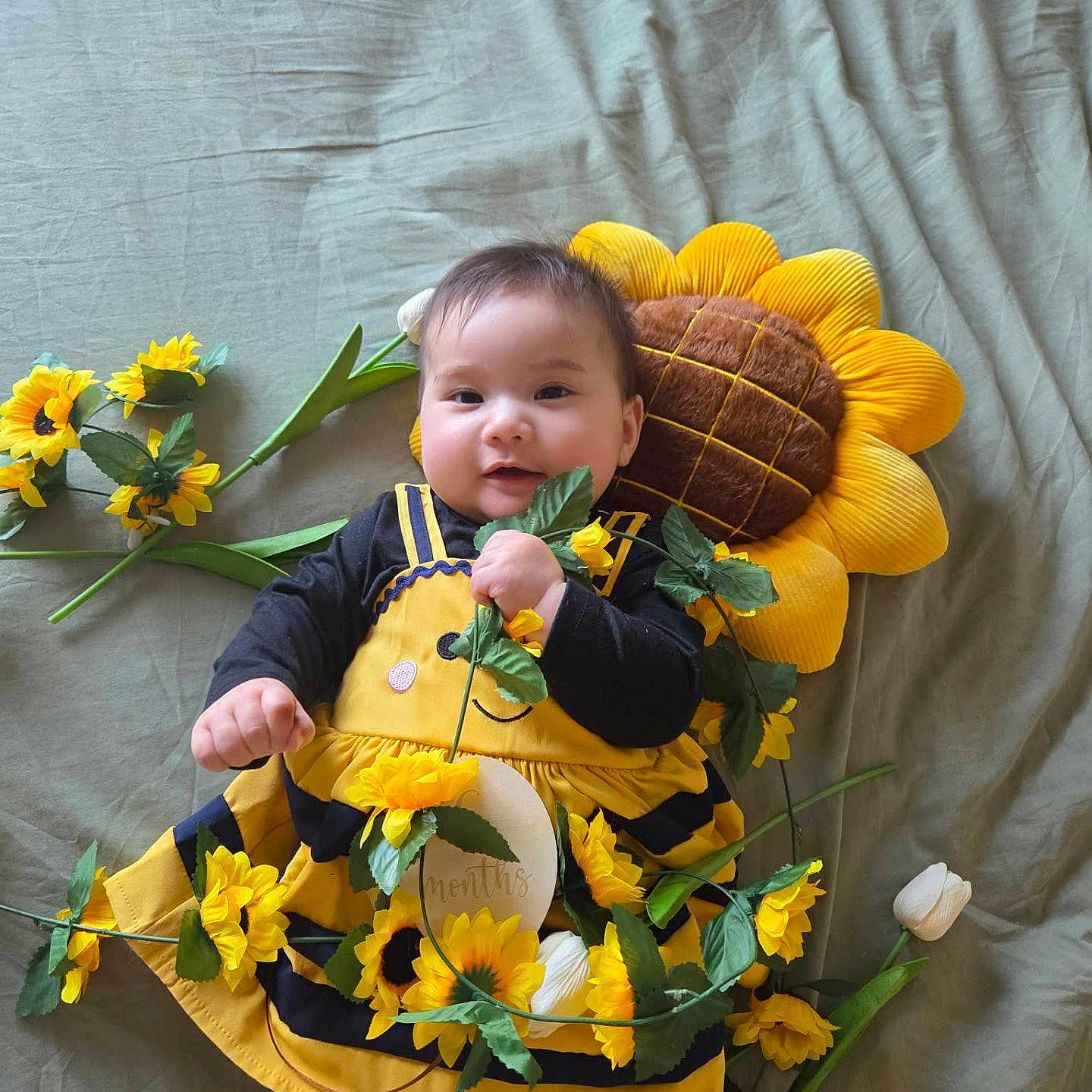 Alena participe au concours pour gagner de l'argent avec cette photo : baby, child, sunflower, plush, flower, yellow, black_clothing, blanket, smiling, cute, lying_down, decorative_flowers, soft_toys, portrait, indoors, infant, holding_object, happy, colorful, playful
