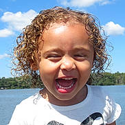 Kyalani is registered to the contest to win money with this photo: child, close_up, clouds, curly_hair, face, happy, lake, landscape, laughing, nature, outdoor, portrait, sky, smiling, summer, sunny, teeth, toddler, water, white_shirt
