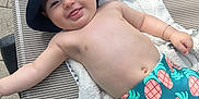 Imrân a rejoint le concours — aidez-le/la à gagner de superbes lots ! baby, smiling, hat, blue_hat, pineapple_shorts, towel, lounge_chair, outdoor, sunny, child, happy, bare_chest, bracelet, tongue_out, playful, relaxing, summer, swimwear, infant, person