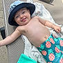 baby, smiling, hat, blue_hat, pineapple_shorts, towel, lounge_chair, outdoor, sunny, child, happy, bare_chest, bracelet, tongue_out, playful, relaxing, summer, swimwear, infant, person