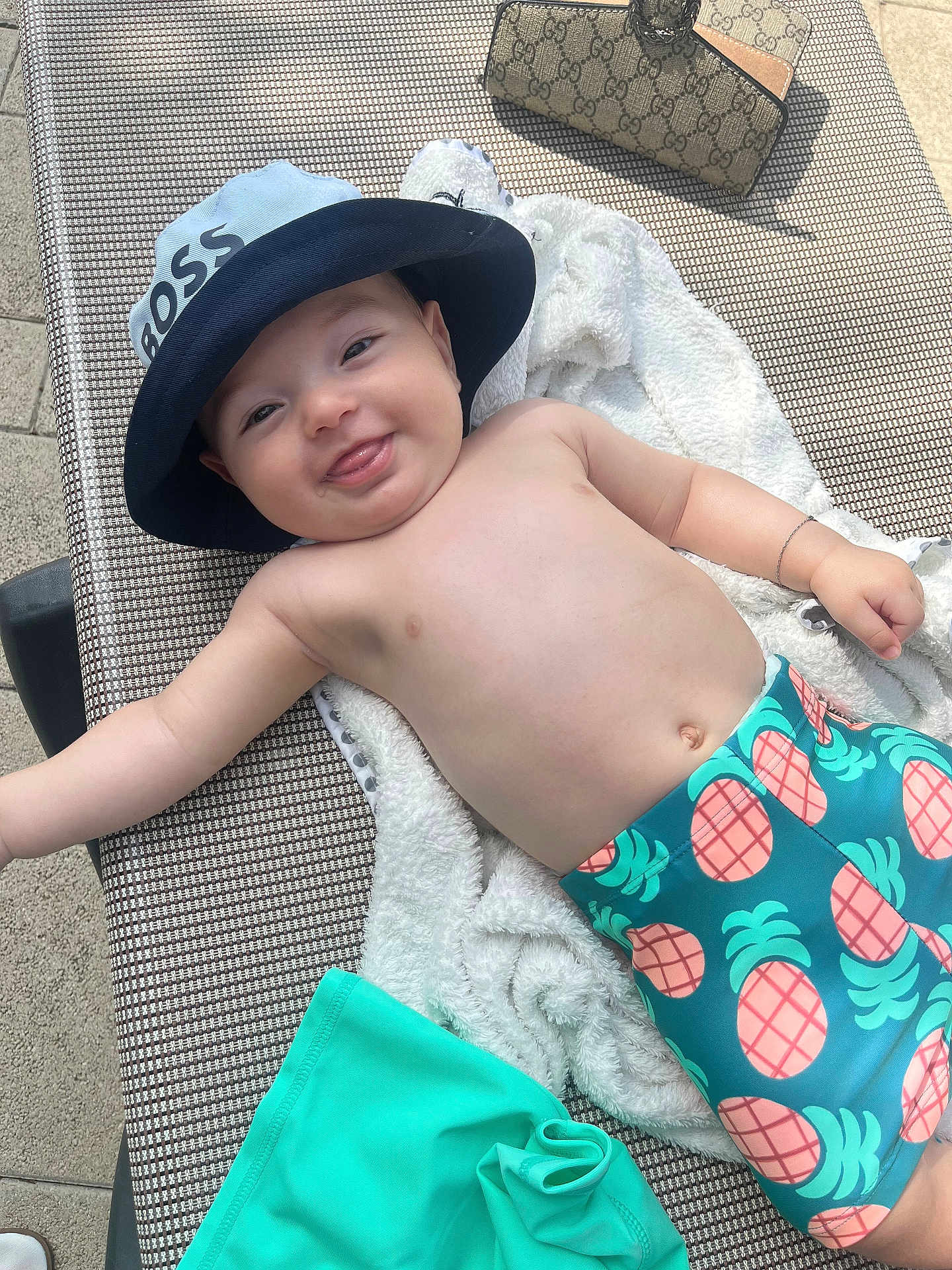 Imrân a rejoint le concours — aidez-le/la à gagner de superbes lots ! baby, smiling, hat, blue_hat, pineapple_shorts, towel, lounge_chair, outdoor, sunny, child, happy, bare_chest, bracelet, tongue_out, playful, relaxing, summer, swimwear, infant, person