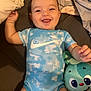 baby, infant, smiling, blue_onesie, nike_logo, stuffed_animal, toy, bed, blanket, happy, face, child, lying_down, cute, indoors, soft_toy, expression, toddler, person, clothing