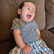 Jorge is registered to the contest to win money with this photo: baby, child, smiling, laughing, couch, clothing, gray_shirt, patterned_pants, indoor, happy, person, seated, infant, cute, young_child, living_room, home, furniture, comfort, expression
