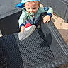 toddler, child, blue_cap, slipper, pacifier, outdoor, wicker_chair, black_chair, sunlight, pavement, baby, casual_clothing, playful, hand, headwear, person, young_child, daylight, hat, reaching
