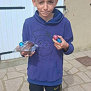 Ethan a rejoint le concours — aidez-le/la à gagner de superbes lots ! child, boy, hoodie, pants, outdoor, pavement, chocolate, candy, playful, winking, standing, casual_clothing, daylight, smile, holding, person, young, blue, yellow, expression