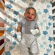 Martin is registered to the contest to win money with this photo: baby, child, infant, smiling, happy, portrait, lying_down, crib, changing_mat, cloud_pattern, mittens, striped_clothing, gray_outfit, nursery, indoor, close_up, face, hands, safe, photography