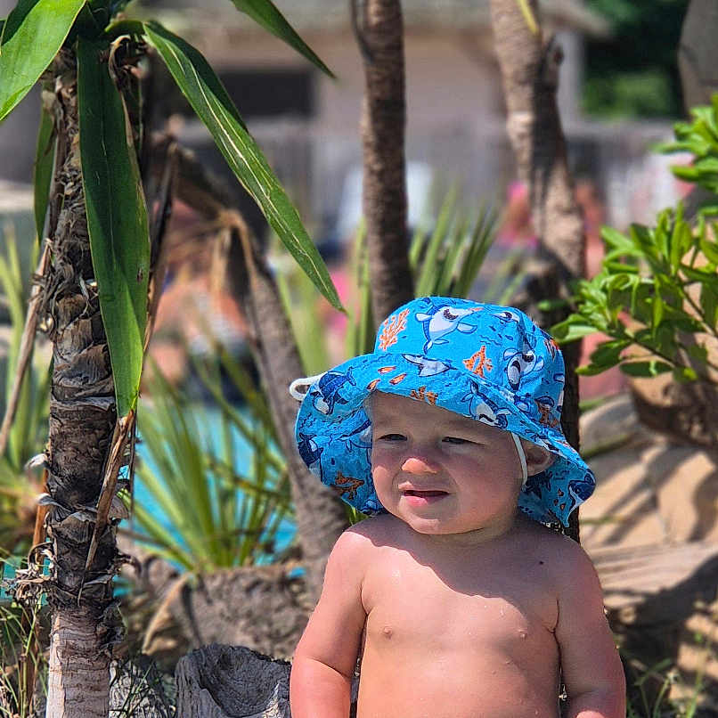 Amaël a rejoint le concours — aidez-le/la à gagner de superbes lots ! barefoot, blue, child, crab_pattern, daytime, hat, nature, outdoor, plants, poolside, rock, shark_pattern, sitting, summer, sunlight, swimwear, toddler, tropical, vacation, water
