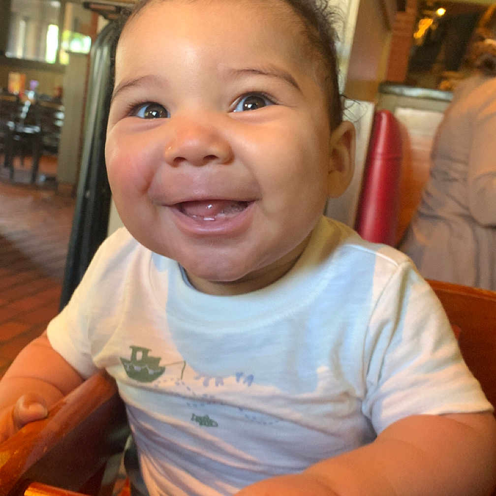 Rakai is registered to the contest to win money with this photo: baby, smiling, high_chair, wooden, indoor, restaurant, child, happy, cute, face, toddler, clothing, seat, person, infant, portrait, joy, skin, hair, hand