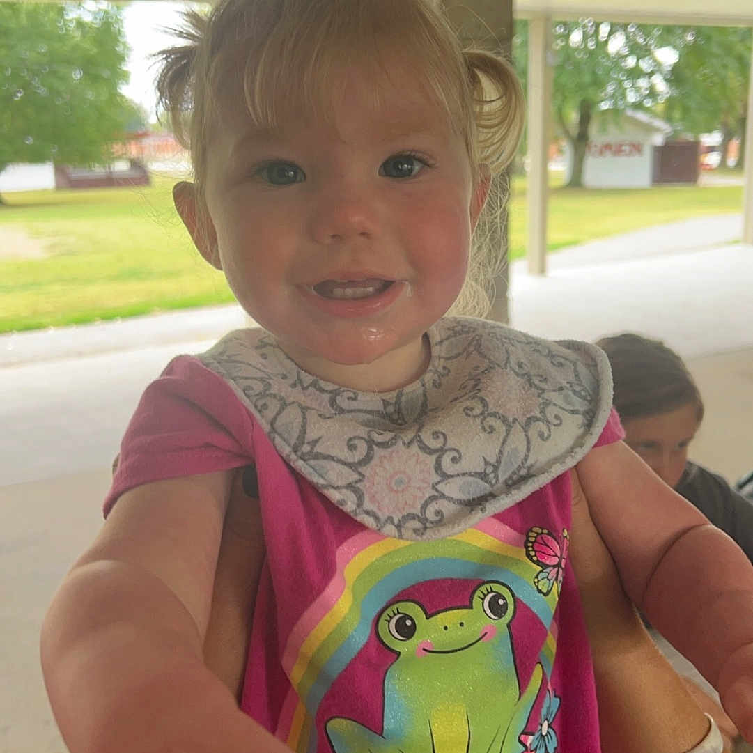 Roxie joined the competition — help win amazing prizes! bib, casual_clothing, child, cute, daylight, frog_graphic, fun, greenery, happy, holding, outdoor, pavilion, person, pink_shirt, playful, portrait, reaching_hand, short_hair, smiling, toddler