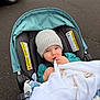 Eli is registered to the contest to win money with this photo: baby, baby_carrier, blanket, bundle, child, cold_weather, curious, cute, hat, infant, knit_hat, outdoor, pavement, person, seat, stroller, teal_clothing, transport, white_blanket, young_child