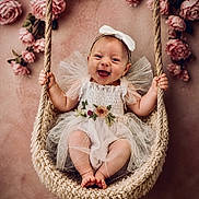 Alaina joined the competition — help win amazing prizes! baby, infant, smiling, happy, swing, knitted_swing, white_dress, bow_headband, floral_embroidery, pink_roses, flowers, flower_background, cute, portrait, child, indoor, decor, soft_lighting, innocence, joy