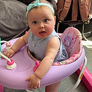 Delilah-Mae is registered to the contest to win money with this photo: baby, child, headband, walker, pink, blue, indoor, floor, clothing, furniture, bag, curious, expression, feet, person, seat, toy, living_room, couch, folded_clothes