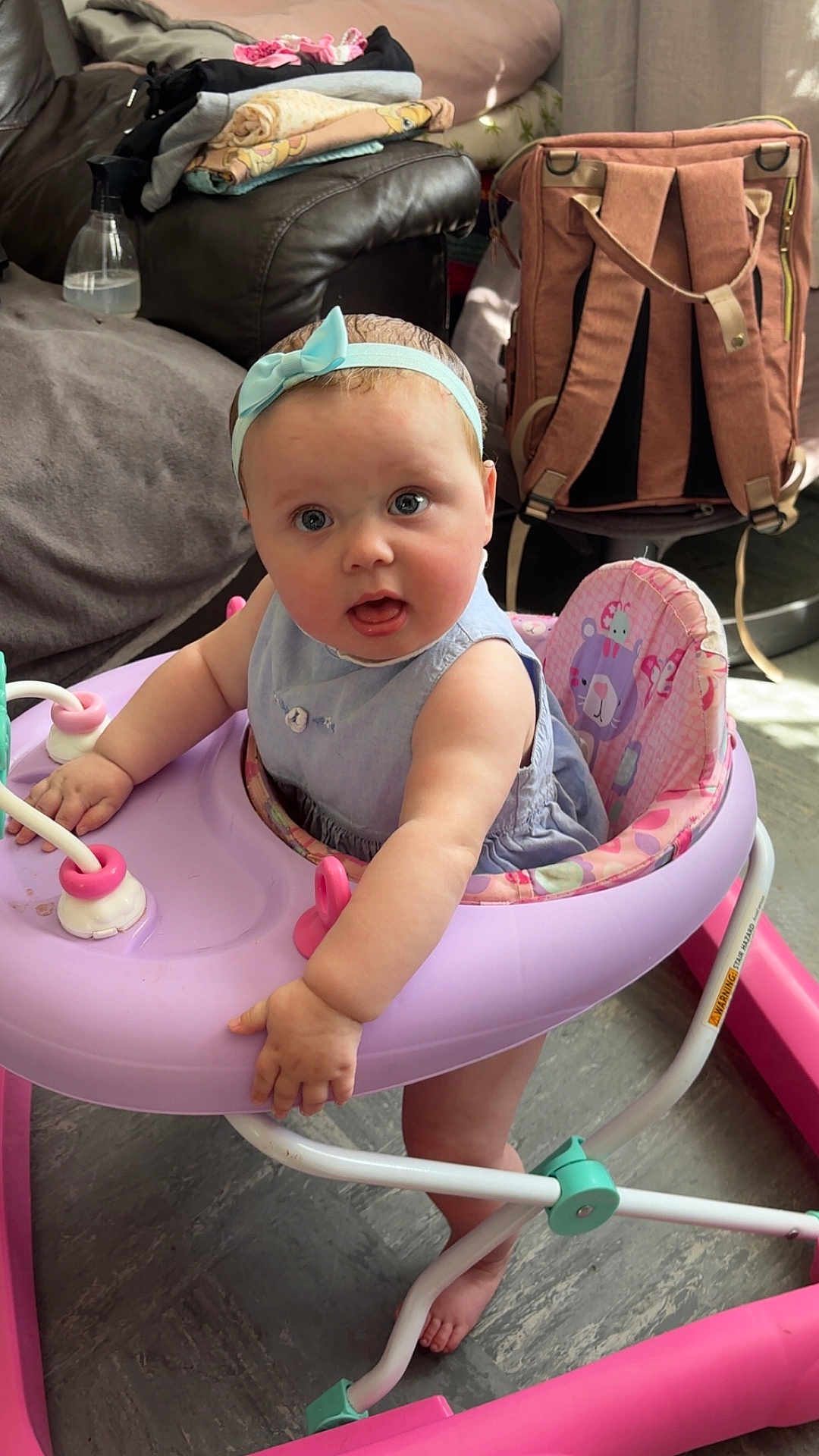 Delilah-Mae is registered to the contest to win money with this photo: baby, child, headband, walker, pink, blue, indoor, floor, clothing, furniture, bag, curious, expression, feet, person, seat, toy, living_room, couch, folded_clothes