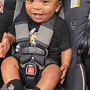 Messiah is registered to the contest to win money with this photo: baby, car_seat, smiling, child, infant, safety_harness, black_clothing, socks, happy, person, seat, indoor, cute, toddler, face, hand, grinning, young_child, clothing, seatbelt