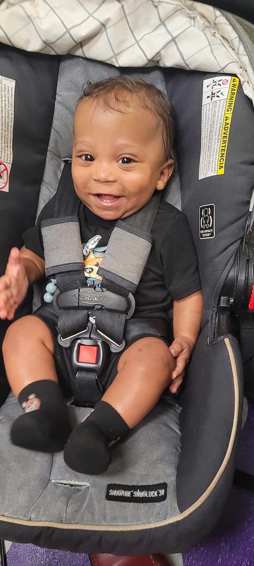 Messiah is registered to the contest to win money with this photo: baby, car_seat, smiling, child, infant, safety_harness, black_clothing, socks, happy, person, seat, indoor, cute, toddler, face, hand, grinning, young_child, clothing, seatbelt