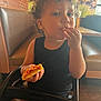 child, toddler, pizza, food, high_chair, curly_hair, black_shirt, indoor, restaurant, table, booth, seat, brown_hair, eating, person, cute, casual, snack, young_child, dining