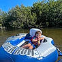 toddler, child, inflatable_raft, water, river, blue_sky, trees, outdoor, nature, summer, curly_hair, life_jacket, relaxing, sunny, daytime, water_bottle, floating, recreation, young_child, leisure